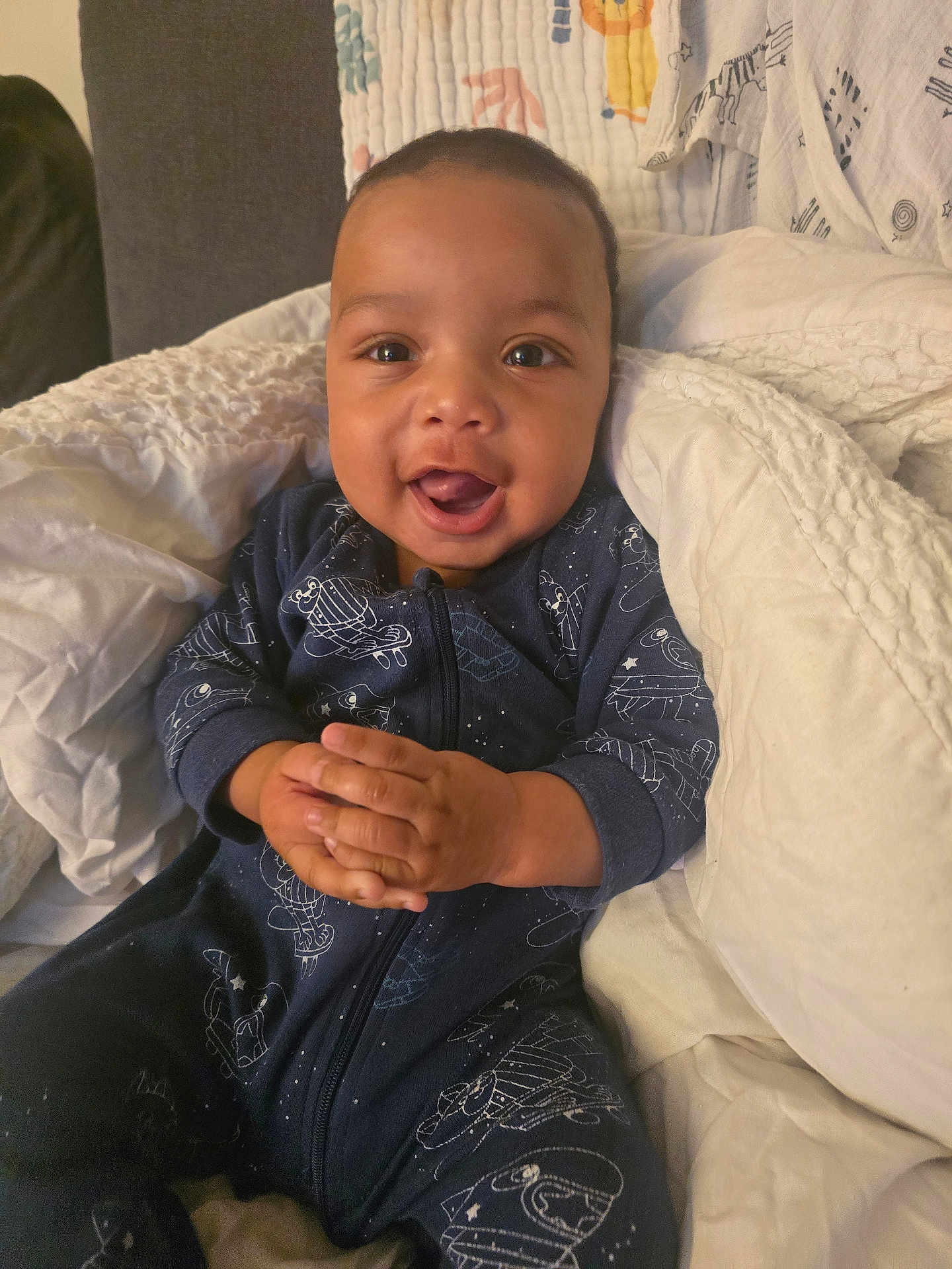 Omari-Momodou is registered to the contest to win money with this photo: baby, child, infant, smiling, clapping, onesie, blue_clothing, blanket, quilt, indoor, cozy, happy, cute, portrait, hands, face, seated, soft_lighting, home, toddler