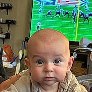 Luka joined the competition — help win amazing prizes! baby, child, infant, portrait, face, eyes, bald, looking_at_camera, serious, indoor, living_room, television, football, sports, football_field, seated, clothing, beige_outfit, baby_gear, home
