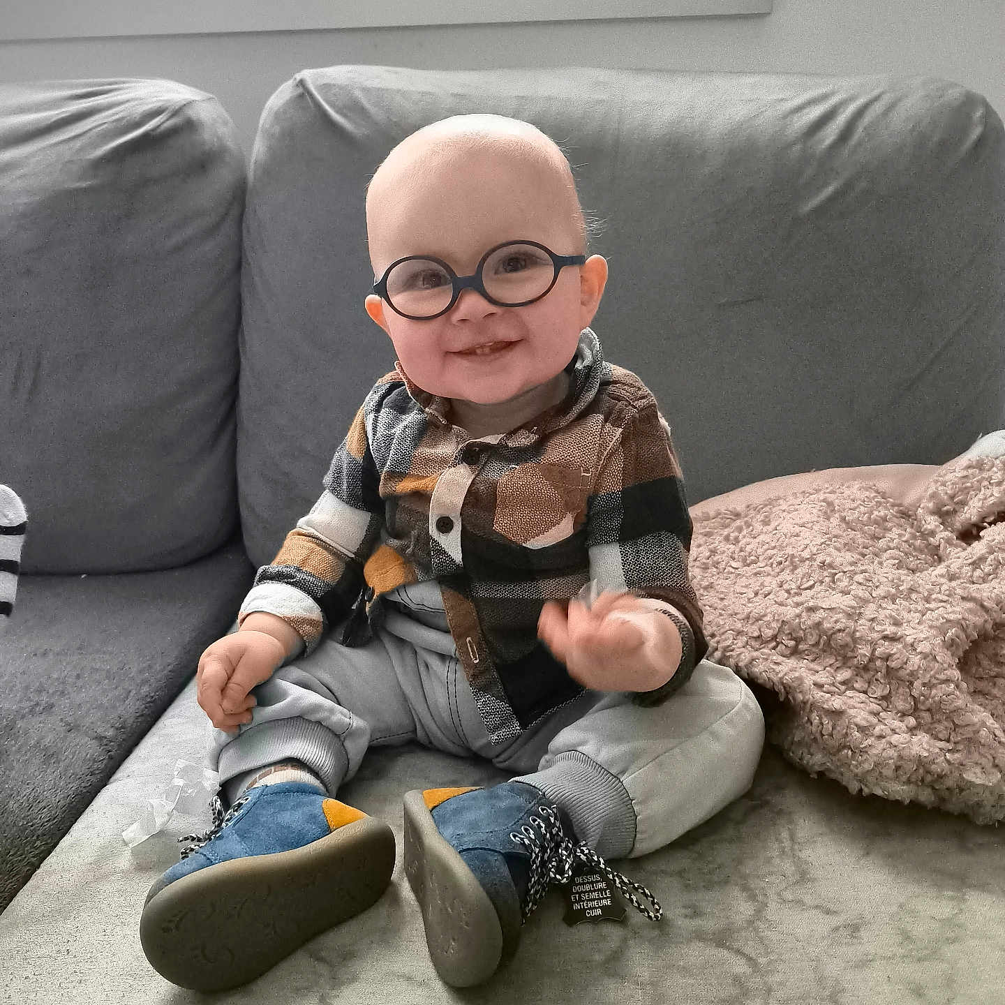 Samael participe au concours pour gagner de l'argent avec cette photo : baby, blanket, blue_shoes, casual_clothing, child, couch, cozy, cute, footwear, furniture, glasses, happy, home, indoor, infant, plaid_shirt, portrait, sitting, smiling, toddler