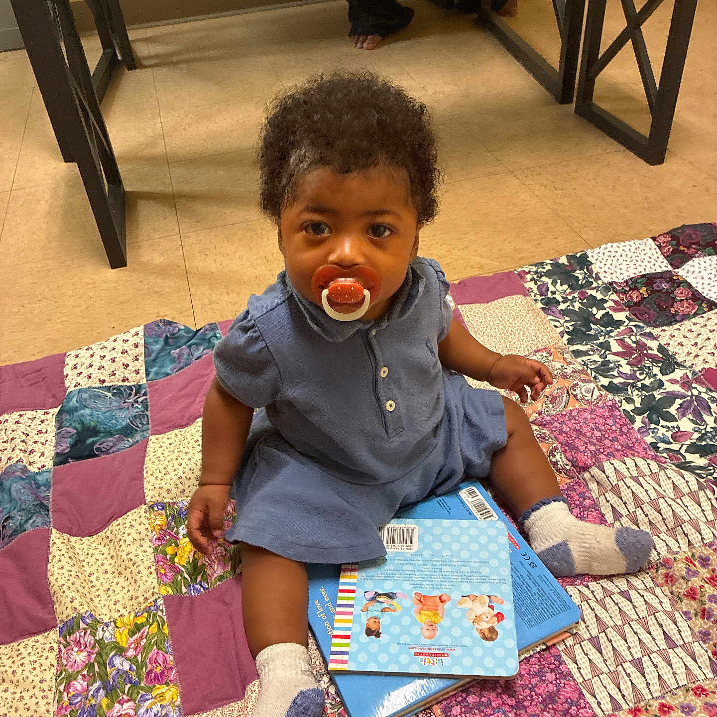 Heaven joined the competition — help win amazing prizes! toddler, pacifier, book, patchwork_quilt, socks, indoor, child, floor, curly_hair, blue_dress, barefoot_adult, wooden_bench, tile_floor, colorful, seated, curious, young_child, cozy, playtime, home