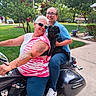 motorcycle, person, dog, outdoor, smiling, glasses, tattoo, casual_clothing, greenery, sidewalk, sunlight, happy, vehicle, leisure, portrait, two_people, pet, short_hair, sneakers, residential_area