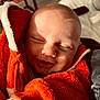 baby, smiling, orange_jacket, fleece, cozy, close_up, face, infant, child, sunlight, shadow, warm, happy, portrait, indoor, cute, soft_texture, clothing, newborn, peaceful