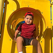 David is registered to the contest to win money with this photo: child, playground, slide, tunnel_slide, yellow, red_shirt, red_cap, backwards_cap, shorts, sneakers, socks, sitting, outdoor, sunlight, blue_sky, portrait, face, playground_equipment, summer, smiling