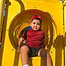 child, playground, slide, tunnel_slide, yellow, red_shirt, red_cap, backwards_cap, shorts, sneakers, socks, sitting, outdoor, sunlight, blue_sky, portrait, face, playground_equipment, summer, smiling