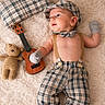 baby, plaid_clothing, hat, mittens, ukulele, crocheted_teddy_bear, pillow, fluffy_rug, infant, cute, child, toy, musical_instrument, relaxed, indoor, portrait, soft_texture, suspenders, barefoot, fashion