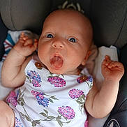 Iris is registered to the contest to win money with this photo: baby, infant, child, onesie, floral_pattern, car_seat, tongue_out, cute, chubby_arms, wide_eyes, expression, portrait, seatbelt, clothing, person, young, skin, hand, face, sitting