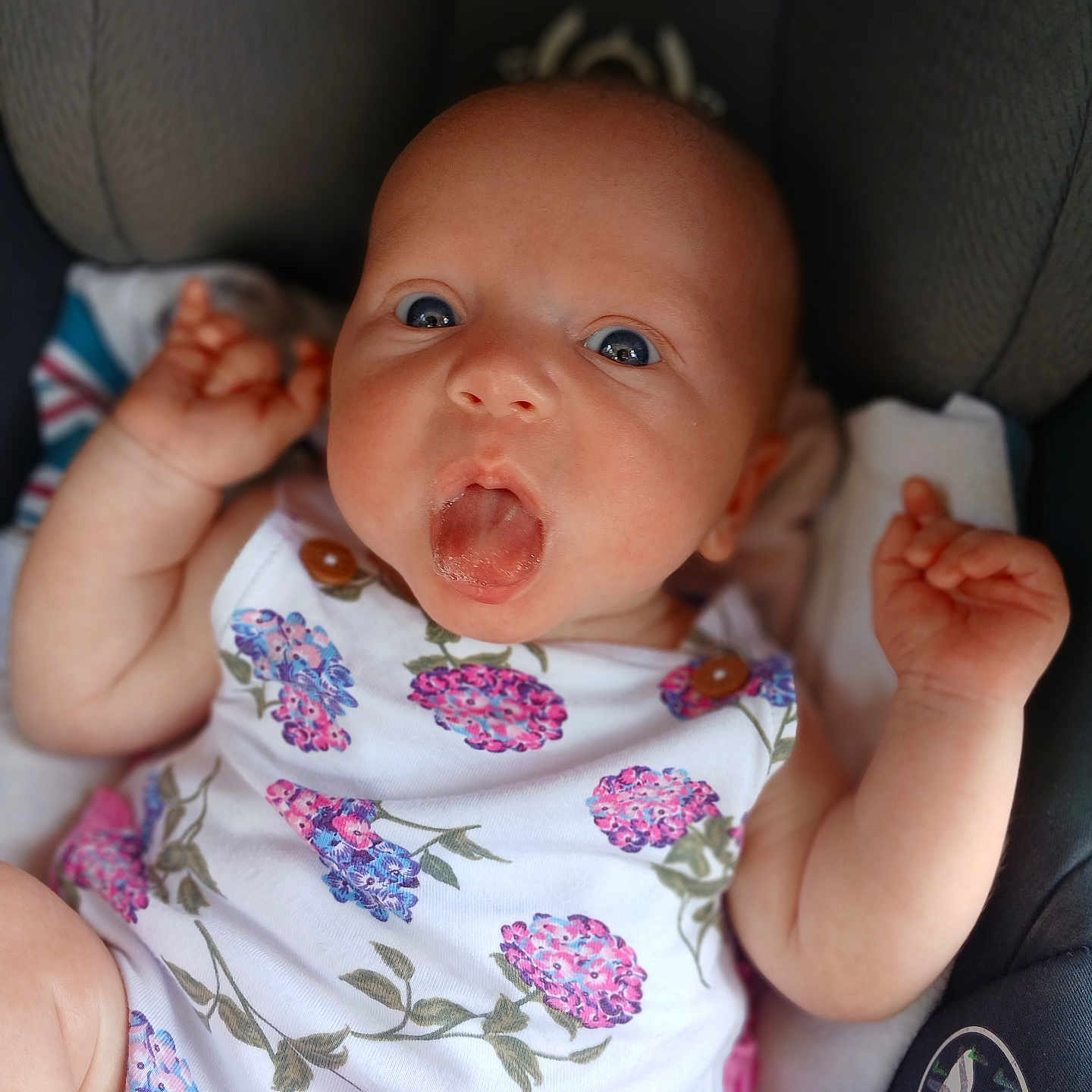 Iris is registered to the contest to win money with this photo: baby, infant, child, onesie, floral_pattern, car_seat, tongue_out, cute, chubby_arms, wide_eyes, expression, portrait, seatbelt, clothing, person, young, skin, hand, face, sitting
