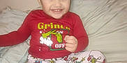 Junior is registered to the contest to win money with this photo: toddler, child, smile, grinch, pajamas, red_shirt, christmas_theme, bed, pillow, indoor, young_child, happy, sitting, cute, holiday, festive, person, face, clothing, home