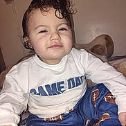 Junior is registered to the contest to win money with this photo: blanket, carpet, child, clothing, cute, expression, face, football_pattern, hair, indoor, light, pants, person, portrait, sitting, smirk, sweatshirt, toddler, toy, wall