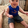 baby, child, infant, sitting, carpet, indoor, portrait, smiling, tongue_out, blue_shirt, shark, shorts, barefoot, adult_leg, home, furniture, cabinet, curtain, cozy, photograph