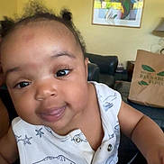 Kamiyah is registered to the contest to win money with this photo: baby, child, infant, face, smile, eyes, clothing, white_outfit, indoor, living_room, lamp, artwork, couch, reusable_bag, person, cute, curly_hair, skin, hand, portrait
