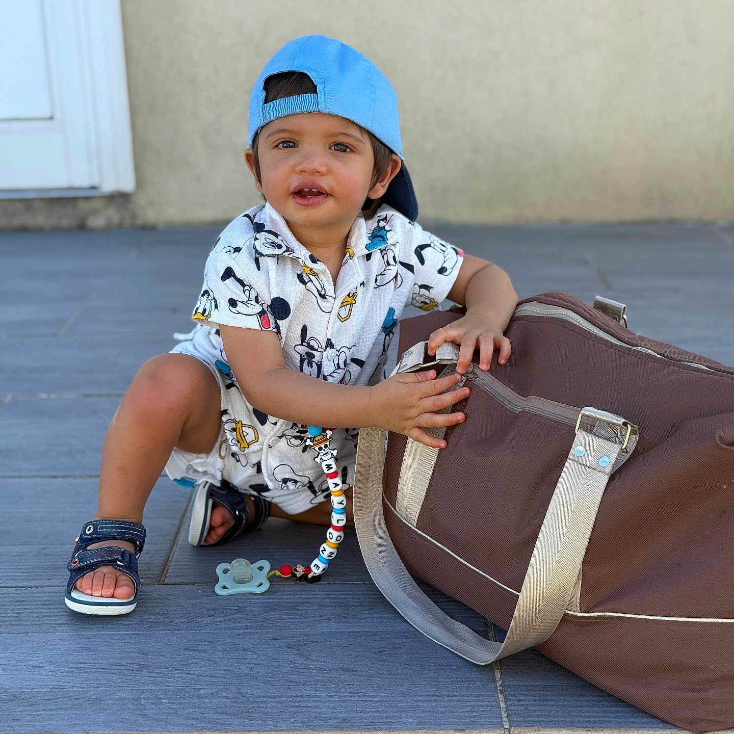 Maylone a rejoint le concours — aidez-le/la à gagner de superbes lots ! blue_cap, child, clothing, curious, cute, daylight, door_frame, duffel_bag, floor_tiles, holding, mickey_mouse_outfit, outdoor, pacifier, person, playful, sandals, sitting, toddler, wall, young_child