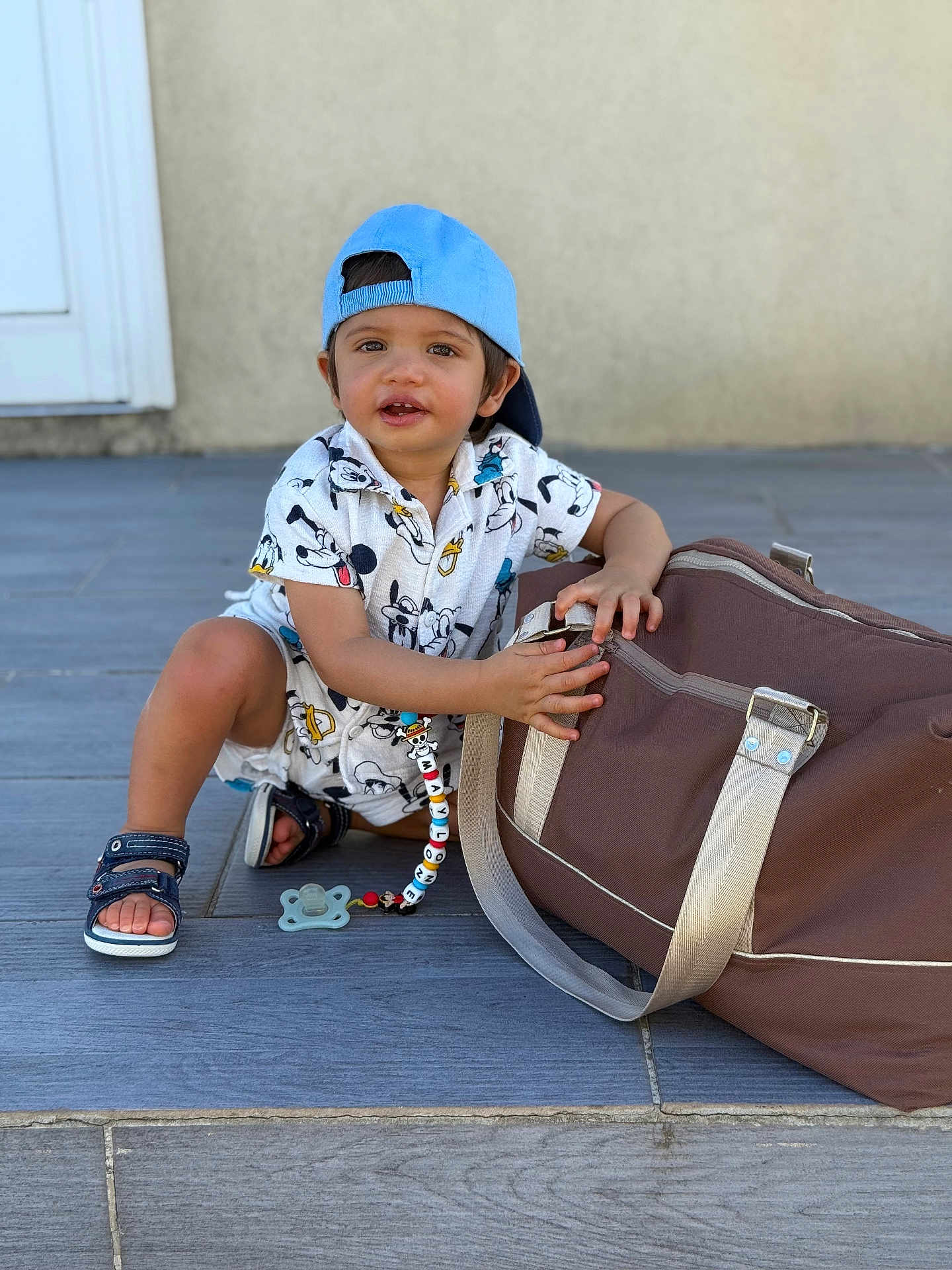 Maylone a rejoint le concours — aidez-le/la à gagner de superbes lots ! toddler, child, blue_cap, mickey_mouse_outfit, sandals, pacifier, duffel_bag, floor_tiles, door_frame, wall, sitting, holding, playful, outdoor, young_child, cute, curious, clothing, person, daylight