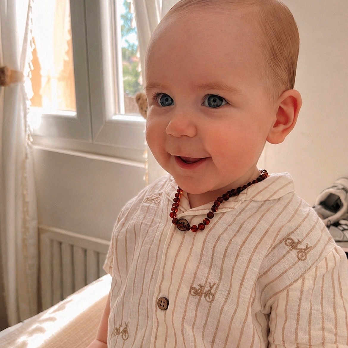 Lyandro participe au concours pour gagner de l'argent avec cette photo : baby, child, indoor, portrait, smile, blue_eyes, striped_shirt, beaded_necklace, window, curtains, natural_light, cute, infant, happy, person, clothing, sitting, face, skin, head