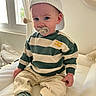 toddler, child, pacifier, cap, striped_sweater, beige_pants, striped_socks, indoor, couch, window, natural_light, curtains, baby, cute, smiling, person, seated, soft_lighting, casual_clothing, portrait