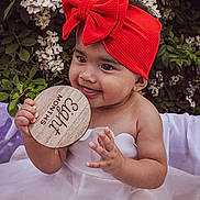 Milani is registered to the contest to win money with this photo: baby, child, red_headband, bow, white_dress, flower, greenery, wooden_sign, eight_months, cute, smiling, portrait, outdoor, nature, skin, infant, hands, sitting, happy, young