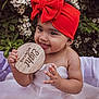baby, child, red_headband, bow, white_dress, flower, greenery, wooden_sign, eight_months, cute, smiling, portrait, outdoor, nature, skin, infant, hands, sitting, happy, young
