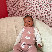 Naomi is registered to the contest to win money with this photo: baby, child, smiling, sitting, pink_outfit, floral_pattern, headband, chair, wallpaper, indoor, portrait, cute, happy, clothing, infant, person, cozy, decor, fashion, texture