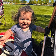 Zakai joined the competition — help win amazing prizes! toddler, child, smiling, outdoor, grass, blanket, curly_hair, sunny, blue_sky, stroller, chair, toy, playing, happy, person, daylight, casual_clothing, reaching_out, park, nature