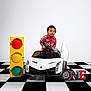 Tishawn is registered to the contest to win money with this photo: child, toddler, toy_car, racing_shirt, traffic_light, checkered_floor, birthday_sign, white_background, indoor, portrait, smiling, cute, car_door_open, decor, studio, happy, black_hair, red_shirt, number_one, photography