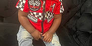 Tishawn is registered to the contest to win money with this photo: toddler, child, sitting, red_shirt, jeans, sneaker, sock, gold_chain, curly_hair, black_couch, indoor, portrait, casual_clothing, looking_away, expression, young_child, fashion, footwear, home_interior, cute