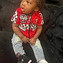 toddler, child, sitting, red_shirt, jeans, sneaker, sock, gold_chain, curly_hair, black_couch, indoor, portrait, casual_clothing, looking_away, expression, young_child, fashion, footwear, home_interior, cute