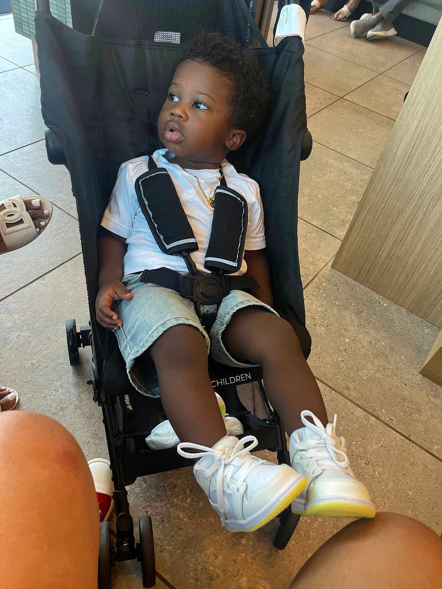 Tishawn is registered to the contest to win money with this photo: toddler, stroller, child, white_tshirt, denim_shorts, white_sneakers, gold_necklace, indoor, tile_floor, curious_expression, casual, seated, footwear, people, legs, black_hair, baby, child_safety, child_seat, person