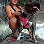 woman, dog, heart_shaped_sunglasses, denim_overalls, pink_harness, graffiti, urban, concrete, leash, lighter, crouching, happy, tongue_out, earrings, tattoo, boots, outdoor, smile, portrait, casual