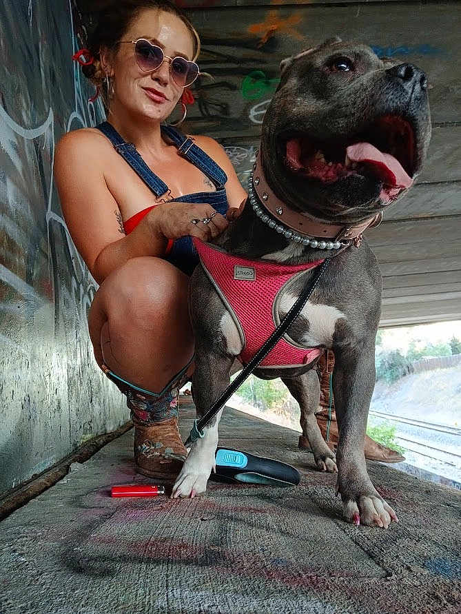 Tesla Marie is registered to the contest to win money with this photo: woman, dog, heart_shaped_sunglasses, denim_overalls, pink_harness, graffiti, urban, concrete, leash, lighter, crouching, happy, tongue_out, earrings, tattoo, boots, outdoor, smile, portrait, casual