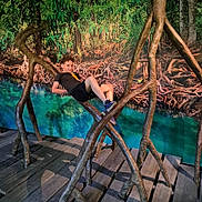 Kenjy a rejoint le concours — aidez-le/la à gagner de superbes lots ! child, boy, curly_hair, wooden_structure, art_installation, roots, indoor, playful, casual, greenery, background, nature_scene, water, turquoise, wooden_floor, shoes, black_shorts, black_tshirt, smile, pose
