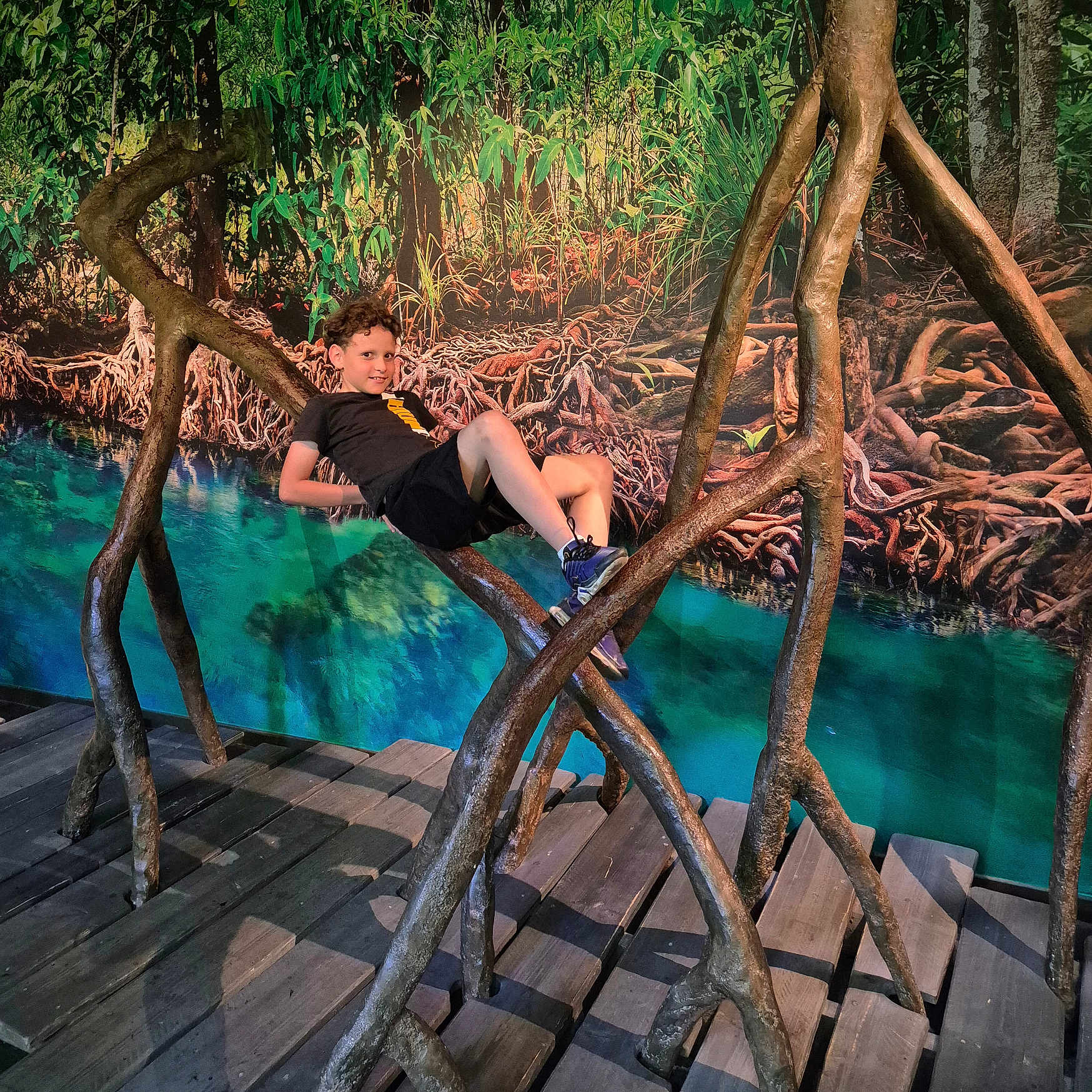 Kenjy a rejoint le concours — aidez-le/la à gagner de superbes lots ! art_installation, background, black_shorts, black_tshirt, boy, casual, child, curly_hair, greenery, indoor, nature_scene, playful, pose, roots, shoes, smile, turquoise, water, wooden_floor, wooden_structure