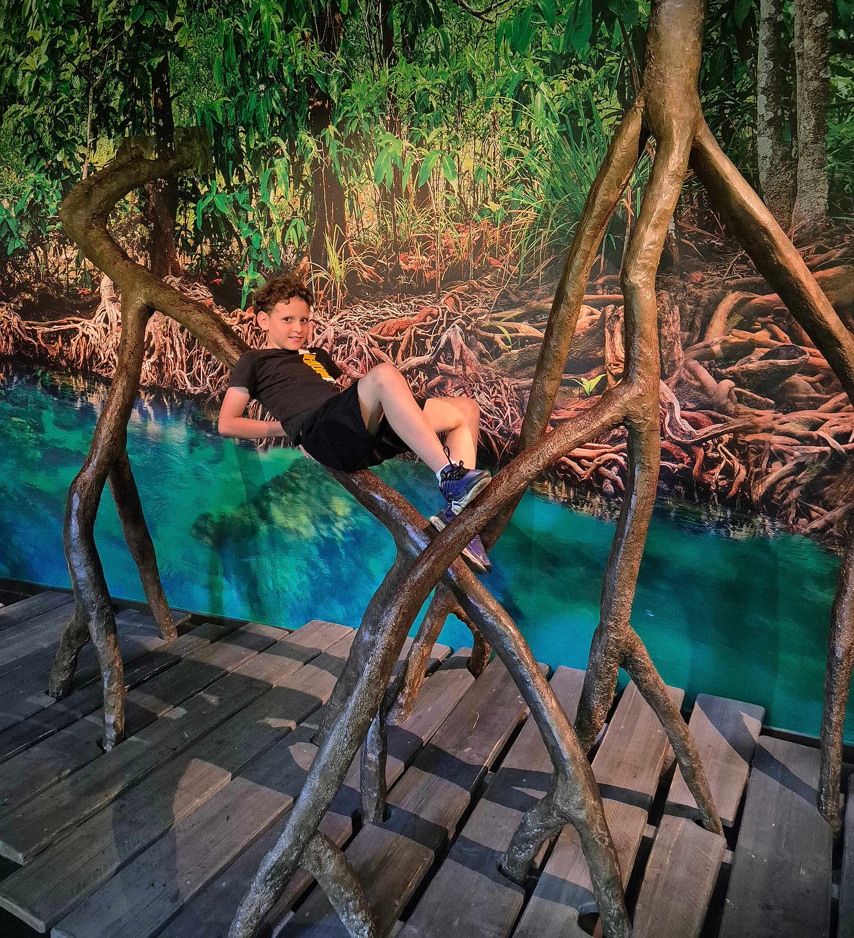 Kenjy a rejoint le concours — aidez-le/la à gagner de superbes lots ! child, boy, curly_hair, wooden_structure, art_installation, roots, indoor, playful, casual, greenery, background, nature_scene, water, turquoise, wooden_floor, shoes, black_shorts, black_tshirt, smile, pose