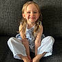 child, smiling, blonde_hair, blue_outfit, striped_clothing, sitting, couch, indoor, happy, portrait, cute, young_child, person, face, barefoot, long_hair, casual, home, light, relaxed