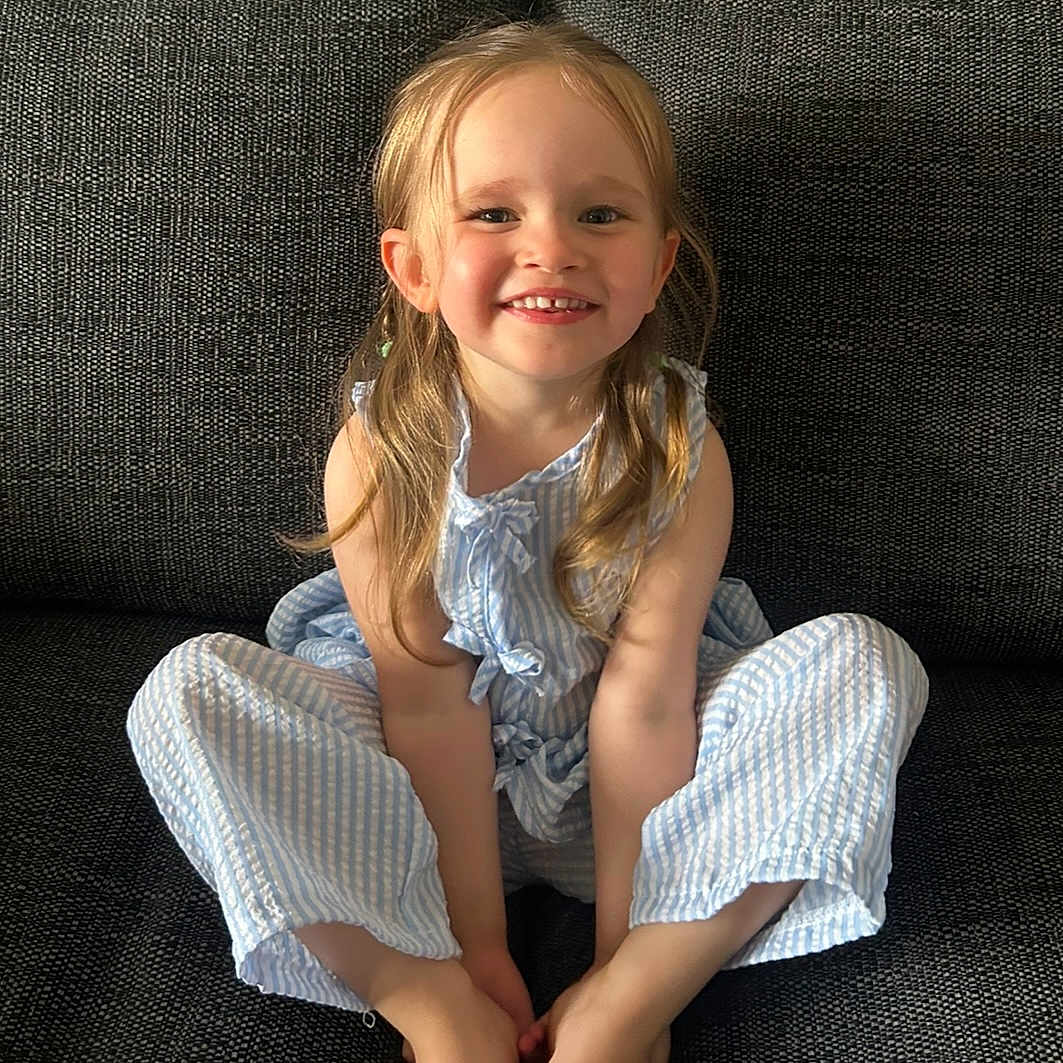 Ava Belle is registered to the contest to win money with this photo: child, smiling, blonde_hair, blue_outfit, striped_clothing, sitting, couch, indoor, happy, portrait, cute, young_child, person, face, barefoot, long_hair, casual, home, light, relaxed