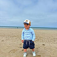 Malo participe au concours pour gagner de l'argent avec cette photo : beach, cap, casual_clothing, child, cloudy, coast, daytime, footwear, ocean, outdoor, person, portrait, sand, shirt, shore, shorts, sky, sneakers, standing, toddler