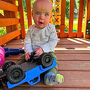Hayden is registered to the contest to win money with this photo: baby, child, toy_car, wooden_deck, play_structure, outdoor, sunlight, curious_expression, sitting, clothing, sock, hand, face, head, playtime, greenery, daylight, wood, blue_toy, casual