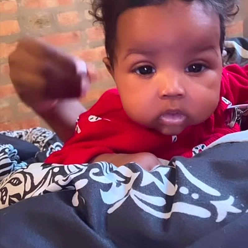 Kahlani joined the competition — help win amazing prizes! baby, blanket, brick_wall, child, closeup, curly_hair, cute, expression, face, fist, indoor, infant, lying_down, motion_blur, pattern, person, portrait, red_clothing, skin, young_child