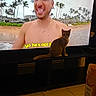 cat, kitten, pet, television, tv_screen, man, shirtless, beach, tropical, palm_trees, ocean, sand, indoor, living_room, furniture, tv_stand, tile_floor, carpet, bottle, subtitles
