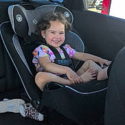 María is registered to the contest to win money with this photo: child, toddler, smiling, car, car_seat, safety_harness, passenger, portrait, indoor, vehicle, outdoor, sunlight, parked_cars, curly_hair, floral_shirt, barefoot, socks, happy, road_trip, family