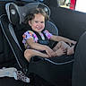 child, toddler, smiling, car, car_seat, safety_harness, passenger, portrait, indoor, vehicle, outdoor, sunlight, parked_cars, curly_hair, floral_shirt, barefoot, socks, happy, road_trip, family
