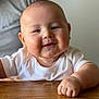Sam is registered to the contest to win money with this photo: arm, baby, child, chubby, closeup, cute, drool, expression, face, hand, happy, indoor, infant, person, portrait, skin, smiling, white_shirt, wooden_table, young
