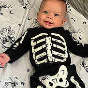 Phoenix is registered to the contest to win money with this photo: baby, child, infant, smiling, portrait, skeleton_costume, halloween, black_clothing, white_bones, bedding, blanket, bed, lying_down, indoors, face, blue_eyes, happy, cartoon_print, spooky, photography