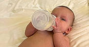 Phoenix is registered to the contest to win money with this photo: baby, infant, bottle, feeding, bed, blanket, bedroom, diaper, lying_down, face, hands, portrait, indoors, home, furniture, sleeping_area, milk, bald, relaxed, parent