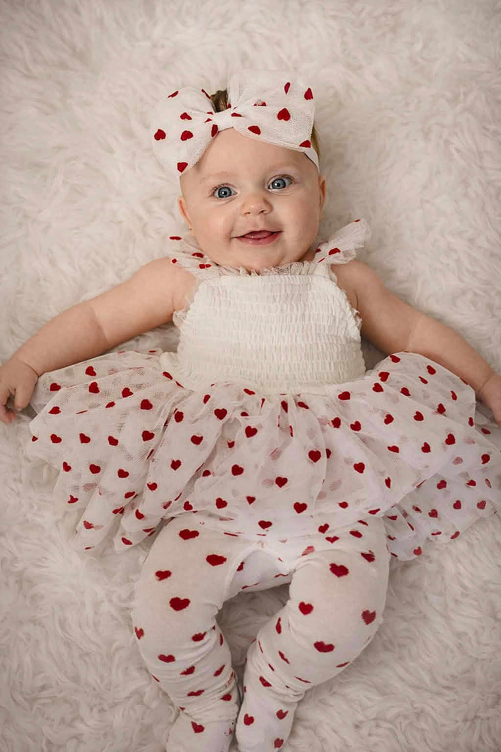 Hazel is registered to the contest to win money with this photo: baby, child, smiling, headband, bow, heart_pattern, white_clothing, leggings, tutu, lying_down, fluffy_background, cute, happy, portrait, infant, blue_eyes, soft_texture, adorable, fashion, playful