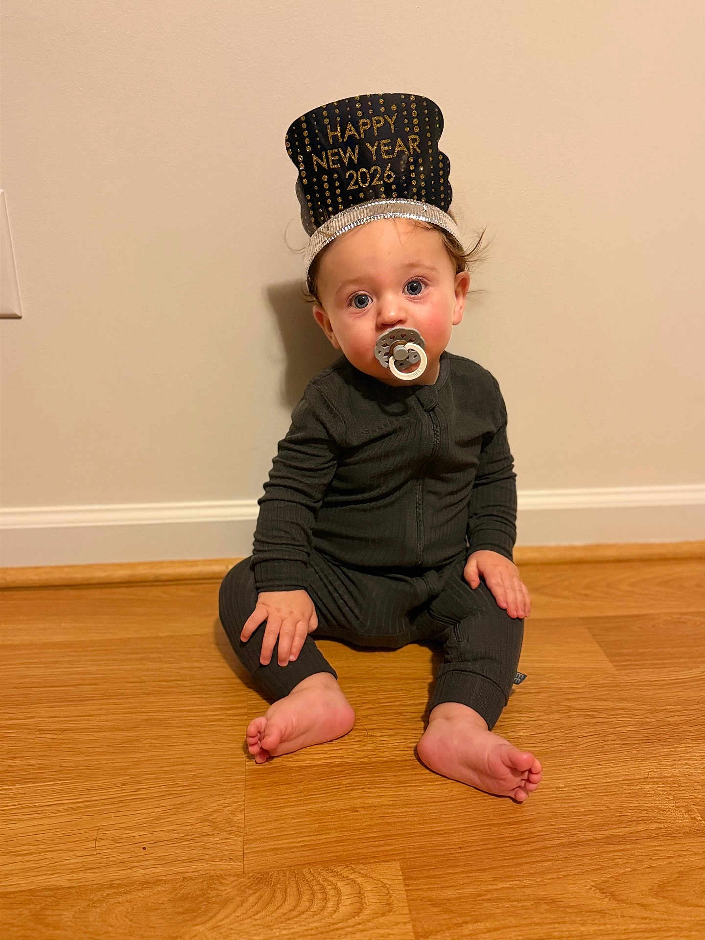 Hudson is registered to the contest to win money with this photo: baby, child, pacifier, headband, happy_new_year, onesie, floor, wooden_floor, indoor, sitting, curious, face, expression, wall, foot, hand, young_child, celebration, holiday, portrait