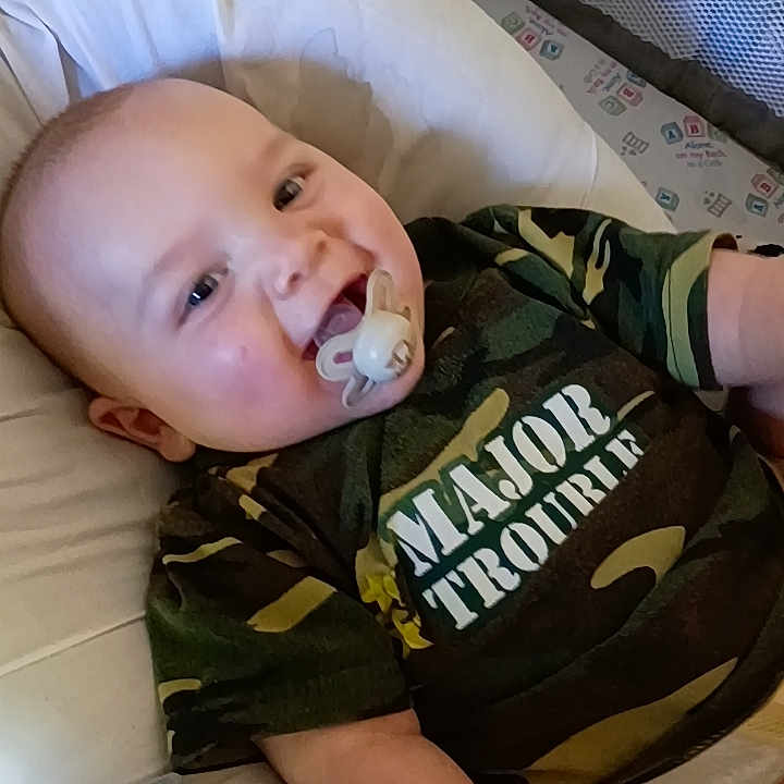 Jax is registered to the contest to win money with this photo: baby, pacifier, camo_shirt, infant, smiling, lying_down, cushion, fabric, indoor, child, cute, casual_clothing, person, young_child, playful, comfortable, relaxed, portrait, human, toddler