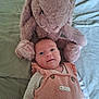 baby, infant, stuffed_animal, bunny, plush_toy, blanket, bed, pink_clothing, smile, face, cute, child, soft_toy, home, resting, lying_down, cozy, happy, indoor, portrait