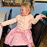 toddler, child, girl, pink_dress, cardigan, chair, black_chair, indoor, smiling, blonde_hair, ponytail, table, tablecloth, wall, stained_glass, furniture, floor, person, happy, portrait