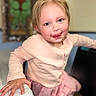 child, toddler, person, face, blue_eyes, pink_clothing, long_sleeve, smile, tongue_out, hand, indoor, table, chair, blurry_background, rosy_cheeks, cute, portrait, happy, young, playful