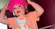 Oakley joined the competition — help win amazing prizes! child, toddler, hat, pink_hat, orange_shirt, cat_graphic, indoor, crib, purple_wall, playful, hands_up, curious_expression, person, cute, smiling, young_child, short_sleeve, toy_hat, home, portrait